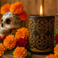 Día de los Muertos candle in eggplant glass tumbler with golden calavera design and matching travel tin with colorful artwork, styled with marigolds for Viviana Luxury.